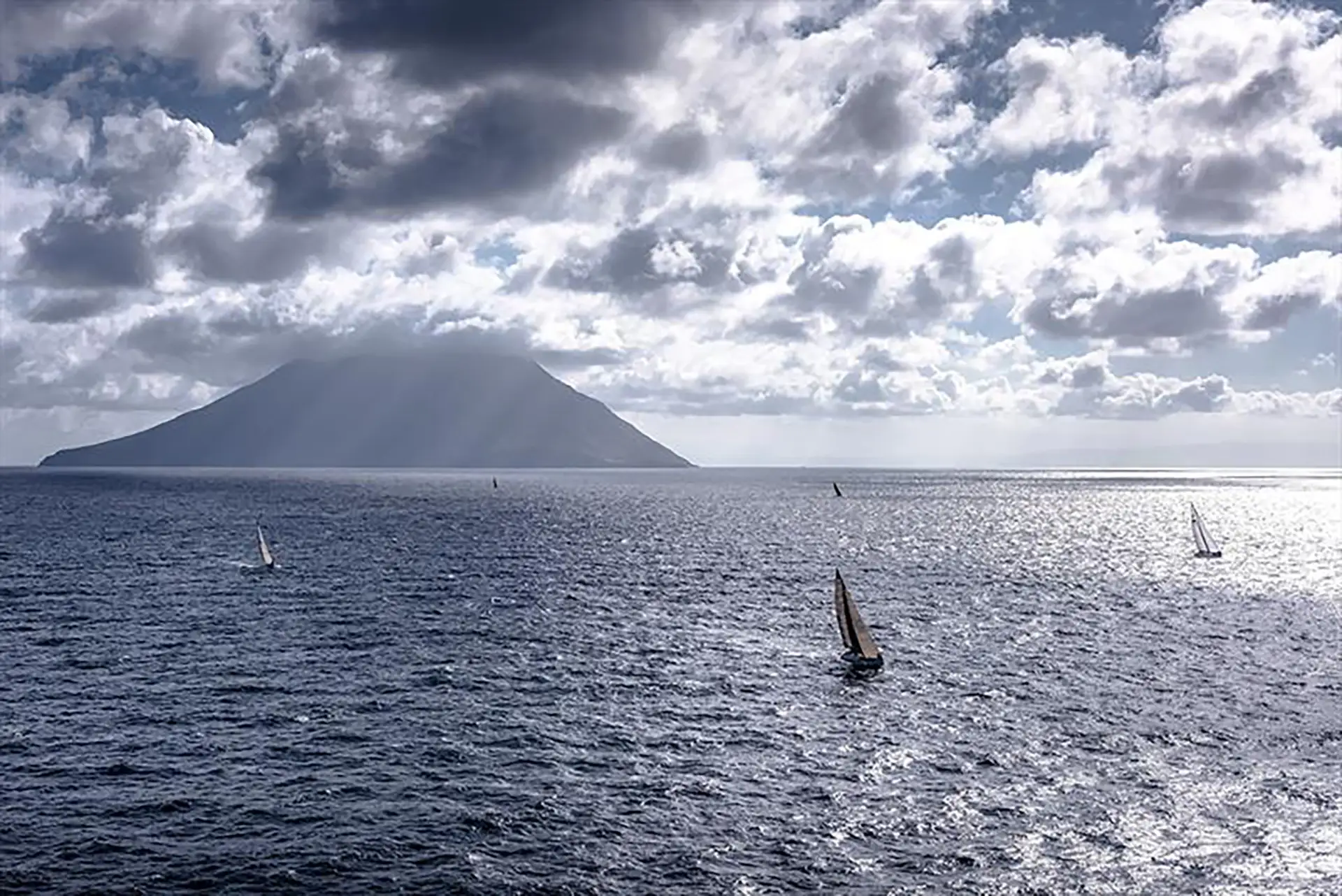 An offshore sailboat race with multiple yachts on a choppy sea. In the distance, the large volcanic cone of Mount Etna is visible, partially obscured by clouds.