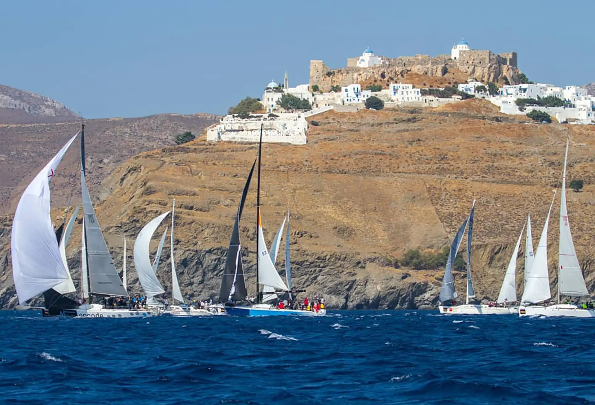 Copyright: Aegean 600/HORC Sailboats racing in the Aegean 600 regatta on deep blue waters, passing below the whitewashed village and Venetian castle of Astypalaia perched on a rocky hilltop.