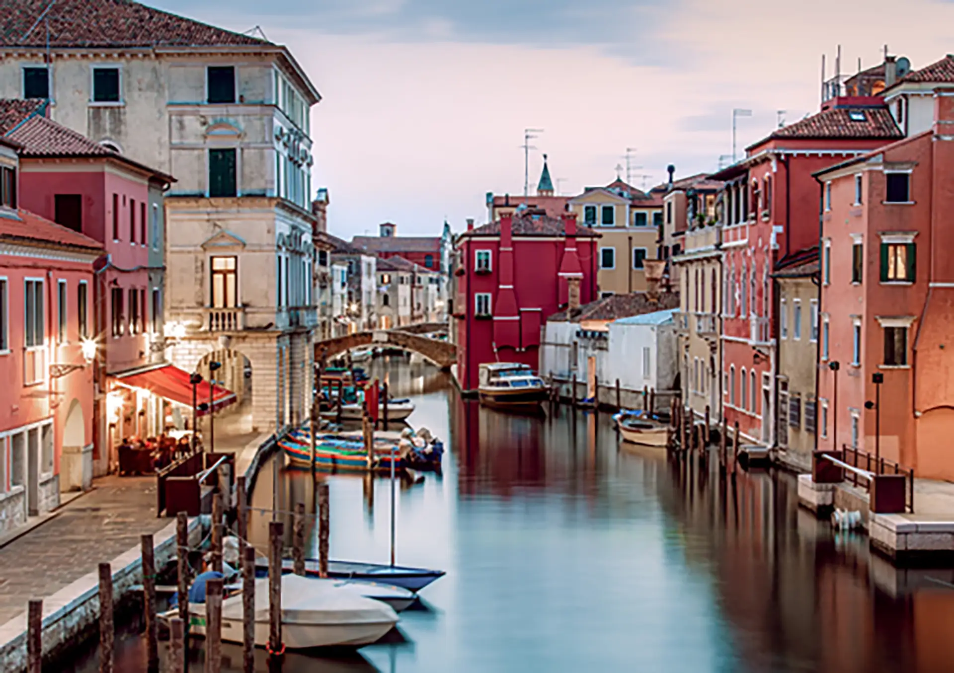 Copyright: COMUNE DI CHIOGGIA, visitchioggia.com A tranquil canal scene in Chioggia, Italy, showcasing the town's vibrant colors and historic architecture. Calm water reflects the pastel-colored buildings lining the canal, with small boats moored along the sides.