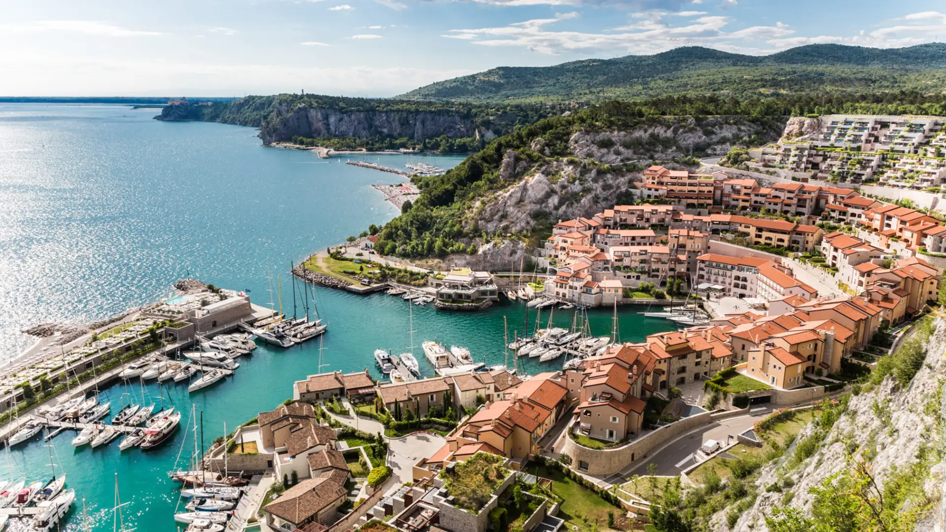 Copyright: Robert Holden Aerial view of Portopiccolo, a modern eco-resort in Italy, blending terracotta-roofed buildings into a cliffside setting overlooking the turquoise Adriatic Sea.