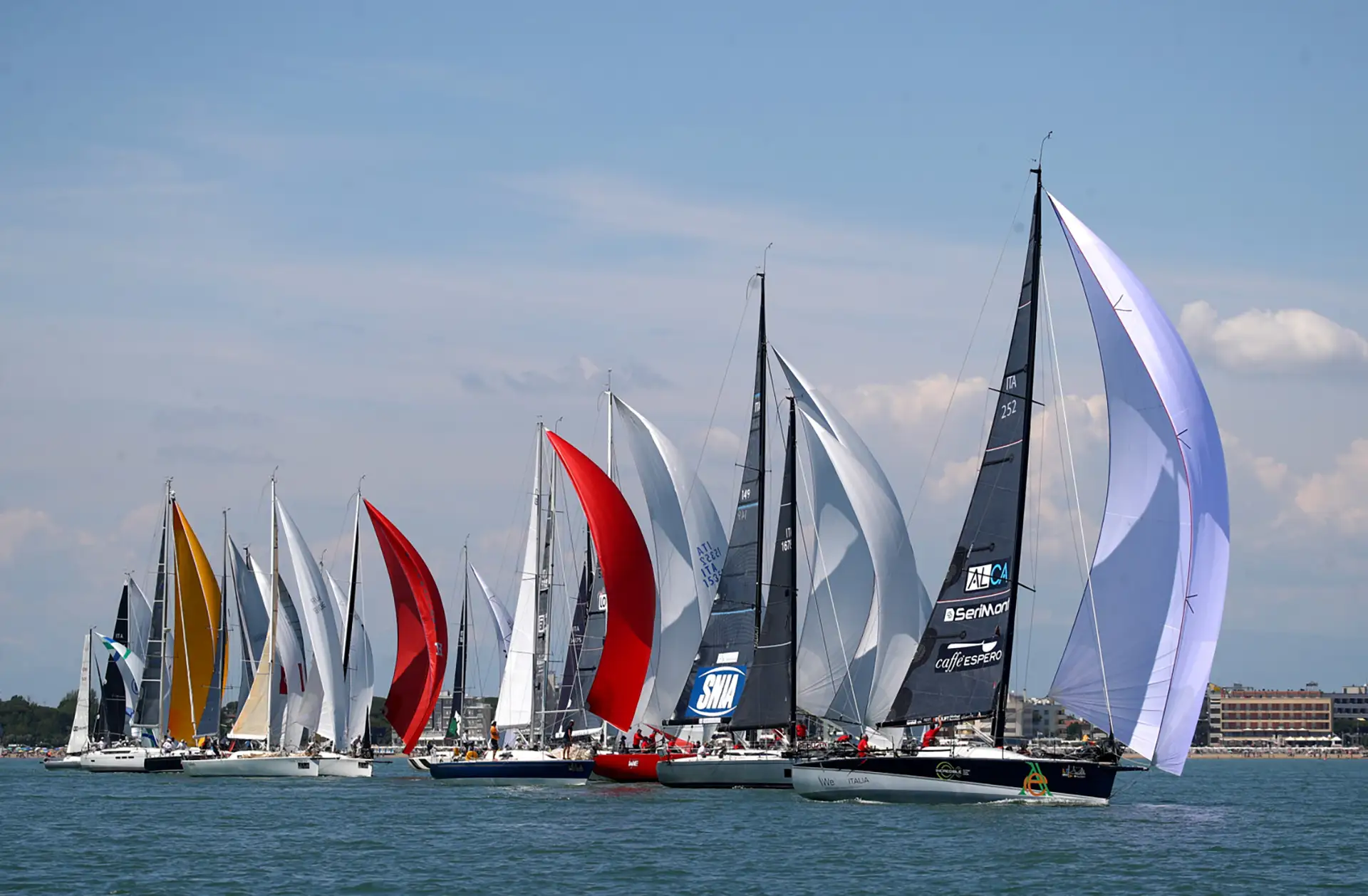 A vibrant sailing regatta featuring numerous sailboats with colorful sails racing on a blue sea under a sky with scattered clouds. The scene captures the excitement and competition of the Circolo Nautico Santa Margherita Caorle.